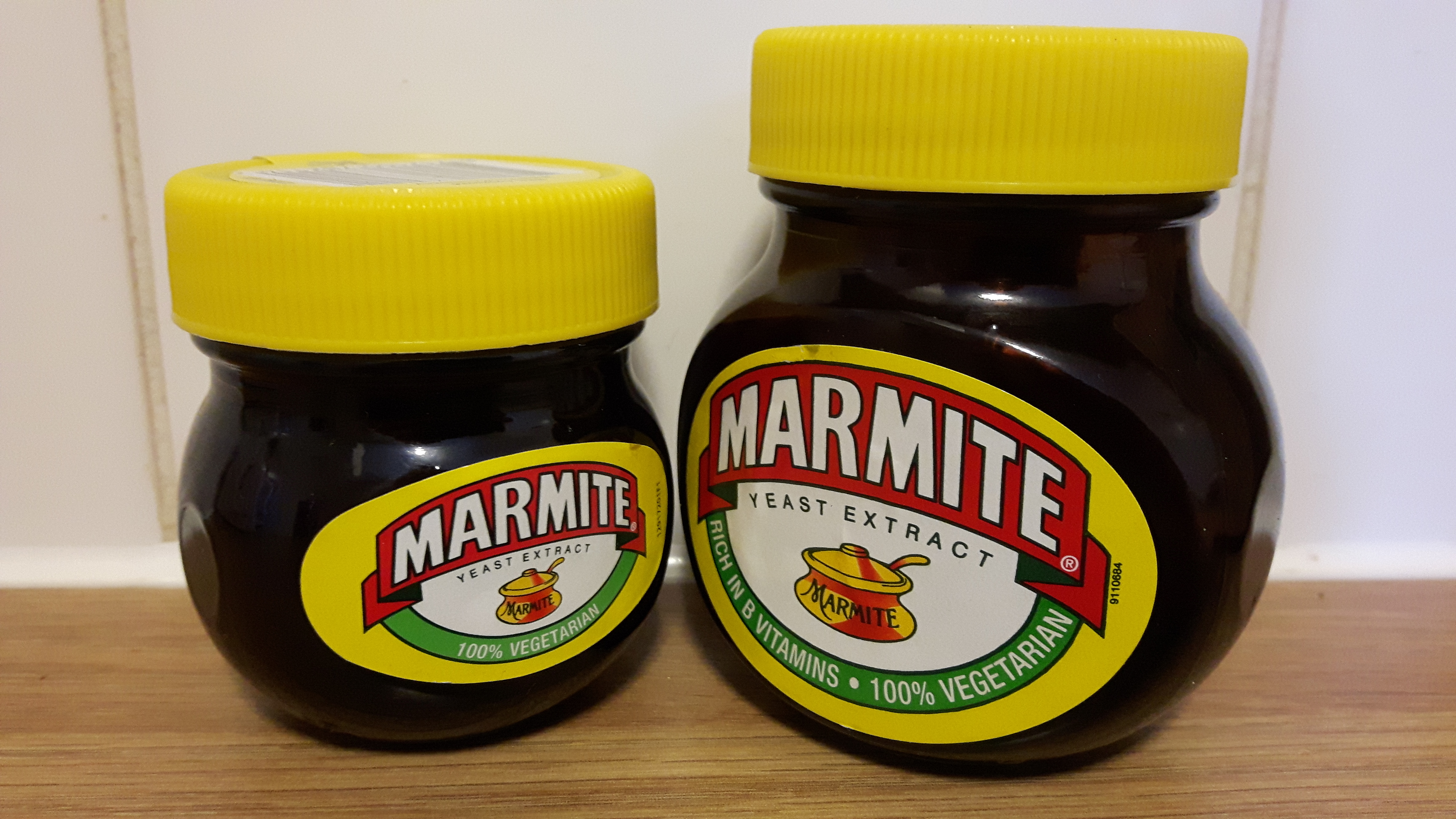 Two jars of marmite. The jar on the left is slightly smaller. They stand on a wooden countertop in front of a white tiled wall. The jars are clean and seemingly unused.